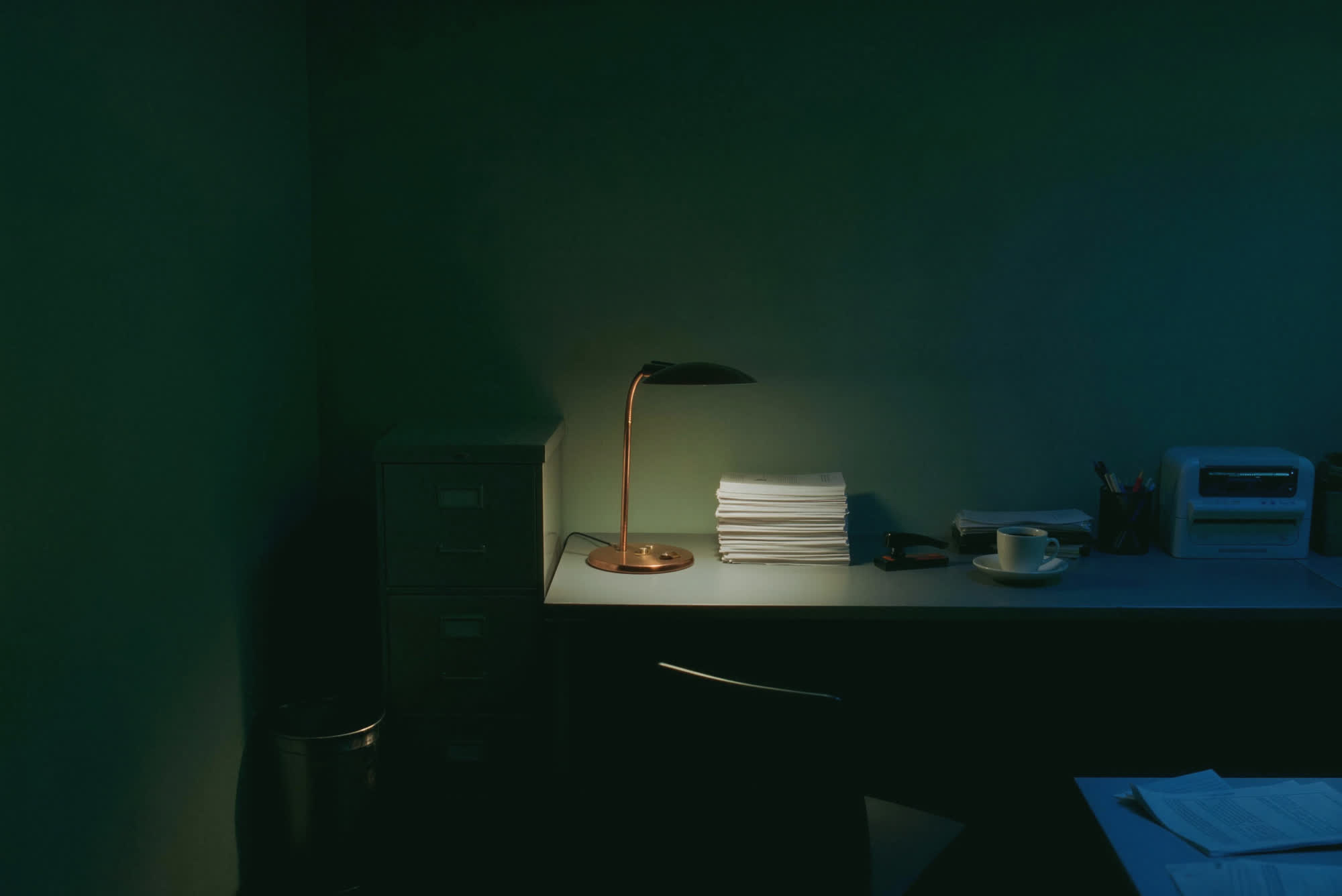 A small clinic back office at night. A copper desk lamp is the only light over stacked day sheets.